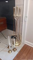 Brass fireplace tool stand with four tools and three brass candle holders with white candles shown on tiled floor near fireplace.