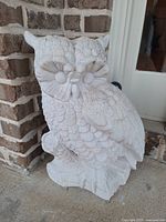 Close-up of hollow concrete owl statue showing detailed feathers and large eyes, white/off-white color, against brick porch background.