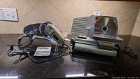 Wide view showing the two meat slicers side by side on a kitchen countertop, with plugs and instruction manual visible.