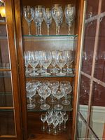 Wooden cabinet shelves with four rows of cut crystal glasses: top row iced tea (12), second row white wine (12), third row champagne (12), bottom row cordial (12).