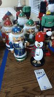 Five wooden nutcrackers with colorful painted details displayed on a wooden surface. Four traditional soldier nutcrackers with crowns and faux fur trims, one penguin nutcracker with a Santa hat and scarf.