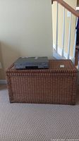 Full front view of the rattan chest with a DVD/VCR player placed on top for scale. Rectangular chest sits on carpeted floor next to staircase.