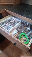 Open kitchen drawer showing various silverware including spoons, knives, forks, serving utensils and green apple slicer