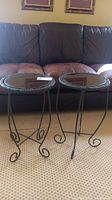 Two small round tables with metal curled legs, rattan trim, and tinted glass tops displayed in front of a brown leather couch.