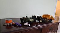 Full view of all vehicles displayed on dresser including military camouflaged Tonka truck, yellow dump truck, tractor trailer, Hot Wheels purple car, Hello Kitty white car, tank vehicle, and black trailer truck