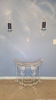 Photo showing the two distressed white metal wire hanging candle holders mounted on wall, each holding a blue pillar candle, and the vintage distressed white wire half-moon stand below on carpet.