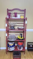 Front view of red Baker Rack style wooden shelf loaded with various decorative and kitchen items for size reference, with tags indicating lot number.