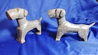Two handmade metal dog sculptures with a rustic iron finish on blue fabric background, showing front and side angles.