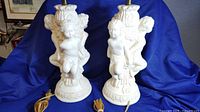 Pair of matching ornate plaster cherub lamps displayed on a blue cloth, showing front view of both lamps including cords and lamp holders