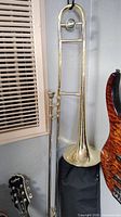 Full view of Accent Brass trombone with mouthpiece, hanging against wall near guitar and window vent.