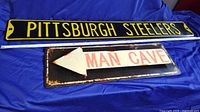 Photo showing both the Pittsburgh Steelers sign and Man Cave directional arrow sign side by side on blue fabric background.