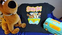 Giant Scooby Doo plush doll sitting next to a Scooby Doo black t-shirt with Mystery Machine design and a Scooby Doo Mystery Machine lunchbox shaped like a van all displayed on blue fabric.