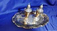 Pair of sterling silver salt and pepper shakers placed on a silver plated tray with ornate scalloped edges on a blue cloth background.
