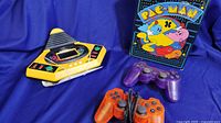 Yellow VTech talking baseball electronic game with buttons and LCD screen alongside purple and orange colored translucent handheld controllers and a Pac-Man puzzle box.