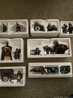 Eight sets of Heritage Village collectible miniature figurines arranged in white foam trays, showing detailed painted village character scenes including carts, horses, stove, and various tradespeople.