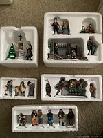 Multiple porcelain Dickens Village figurines shown inside styrofoam packaging, includes a street lamp scene, various carolers, a Victorian gate scene and horse sleigh.
