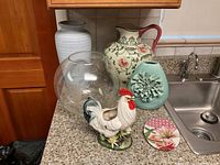 Photo showing a white ribbed ceramic vase, floral ceramic pitcher, clear glass vase, teal flowered vase, ceramic rooster figurine, and floral decor piece on a countertop.