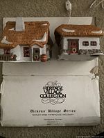 Two white and brown porcelain village buildings with snow accents on the roof, displayed on carpet with the original box underneath showing the Heritage Village Collection branding.