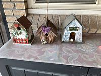 Three decorative birdhouses placed on a table outside against a brick wall. Left is white with red heart and fence, middle rustic wood with purple flower, right gray with floral design and sign.