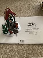 Photo of porcelain figurine showing a gazebo with children figurines ringing bells and a snow-covered Christmas tree. Also shows included Heritage Village Collection informational sheet with product details.