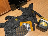 Two black Colorado Buffaloes rain ponchos, three tablecloths (mustard yellow, black and white checkered, brown and black plaid) laid on floor.