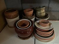 Photo showing a collection of flower pots of various sizes and colors stacked and arranged together on a white surface against a cinder block wall.