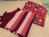 Two hand knitted afghans shown folded on a bed. One afghan is made of colorful granny squares and the other is striped in shades of red and pink.