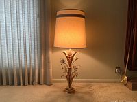 Full view of the tall table lamp lit, showing beige shade with gold trim, gold and white floral base, placed on carpeted floor adjacent to window with white curtains.