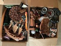 Top view of two open cardboard boxes filled with miscellaneous small decorative items including wood carvings, a model ship, ceramics, and metal pieces.