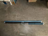 Folded portable movie screen in blue metal casing with disassembled metal stand rods beside it, placed on concrete floor.