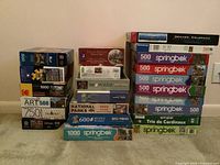 Photo showing 18 puzzle boxes stacked in three columns, brands include Springbok, Kodak, National Parks, and others, varying between 500 to 1000 pieces.