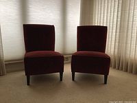 Two red velvet side chairs side by side on carpeted floor with window blinds in background