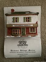 Front view of handpainted porcelain "Scrooge and Marley Counting House" building with detailed shutters, windows and snow accents on roof and sill. Building attached to original display backing with brand and series info.