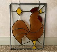 Stained glass wall hanging depicting a brown chicken with red and yellow stained glass pieces in a rectangular leaded frame. Hanging hook visible.