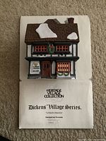 Front view of the handpainted porcelain Tutbury Printer building with visible snow patches on roof, detailed windows, and door, on paper backing labeled 'Heritage Village Collection Dickens Village Series Tutbury Printer'.