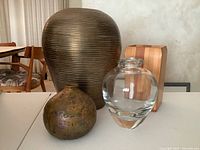 Photo of three vessels on table: large ribbed brass vase, round rustic metallic vase, and clear thick glass vase.
