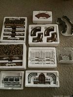 Multiple miniature village accessory pieces laid out inside Styrofoam packaging, showing brown stone walls, white fences, and curved walls.
