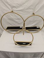 Three brass-colored circular wall shelves in small, medium, and large sizes placed on white surface against white fabric background. Each has black rectangular shelf inside the round frame.