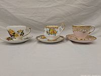 Three Coalport teacups with matching saucers, various floral patterns with gold trim, arranged side by side on a light background.