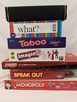 Stack of six boxed board games including What?, Taboo, Imaginiff, Scrabble, Speak Out, Monopoly