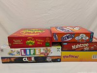 Photo showing six boxed family board games stacked: Apples to Apples Party Box, Ikewise, Yahtzee, Pie Face, Life, and Clue.