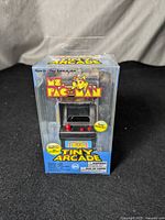 Front view of the Ms. Pac-Man Tiny Arcade game inside its original clear plastic box showing the miniature arcade cabinet with joystick and button controls.