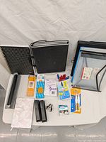 Top view of office supplies including multiple binders, pencils, pens, staplers, notepads and tape dispenser on a white surface.