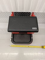 Top view of the black vinyl padded seat with Uline logo and red frame base with visible caster wheels and pullout drawer underneath, tape measure on floor for scale.