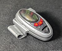 Close-up photo of the silver Bandai Chrono Morpher with red buttons and markings, placed on a dark textured surface.