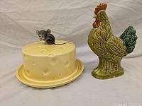 Side view of ceramic yellow cheese covered plate with small mouse figurine on top and green ceramic rooster figurine standing beside it.