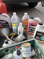Multiple bottles of automotive fluids including Prestone RV WaterLine antifreeze, motor oil, windshield washer fluid, and others with a metal funnel and oil siphon in a green crate