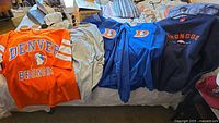 Five pieces of men's Denver Broncos apparel laid out on a bed, showing a bright orange throwback t-shirt, grey t-shirt, two blue pullovers, and a navy sweatshirt.