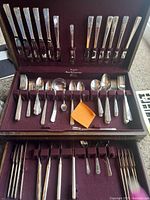 Full view of opened flatware case showing arranged knives, forks, spoons, and serving utensils on purple velvet lining.