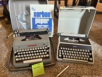 Both Royal typewriters side-by-side in their gray hard cases with accessories including carbon paper and manuals visible.